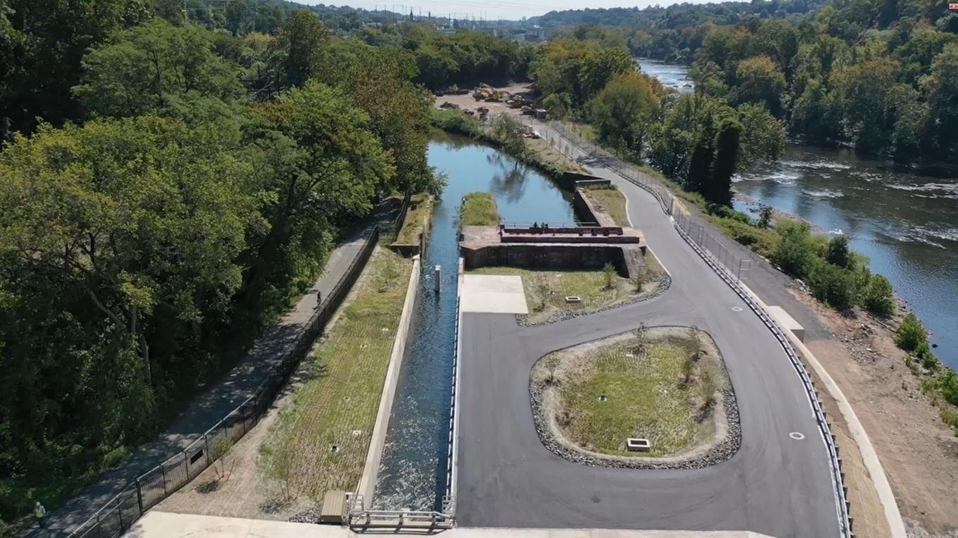 Aerial view of a restored canal lock and adjacent river, showing a narrow water channel, paved access road, landscaped embankments, and surrounding green woodland.