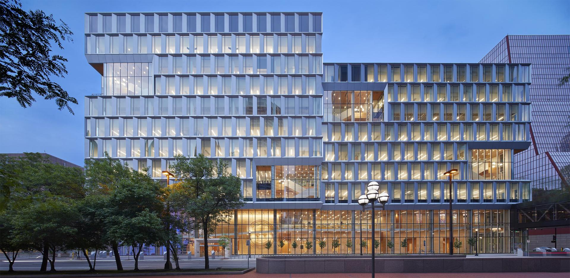 A modern, multi-story glass building at dusk with a facade made of stacked, rectangular sections. Warm light glows from the large windows, and leafy green trees line the sidewalk in the foreground.